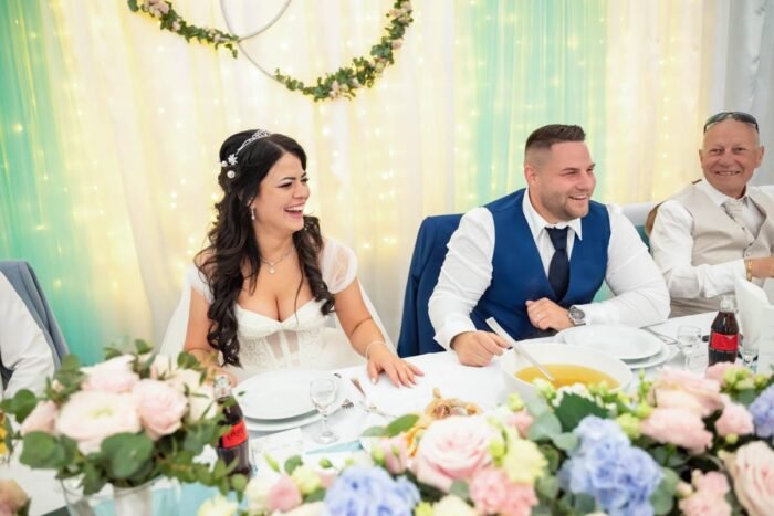 Az esküvői vacsora közben a boldog pár és a vendégek vidáman nevetnek, miközben az asztalon étkezési kellékek és friss virágok díszítik a terítéket. Az ifjú pár örömteli pillanatait az asztal körül ülők is osztoznak, ami még varázslatosabbá teszi az eseményt.