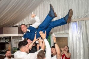 A tánctér közepén a vőlegény szárnyal a levegőben, miközben a többiek ünneplik őt. A felemelt férfi hatalmas mosollyal és nevetéssel élvezi a pillanatot, míg a társai az égből emelik, miközben a háttérben a táncoló vendégek is jól szórakoznak a pörgős, szórakoztató buli során. A tánc és a jó hangulat a legjobb pillanatokban csúcsosodik ki.
