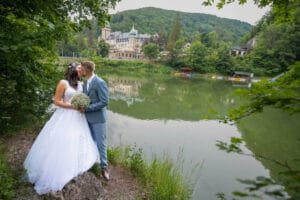 A menyasszony és a vőlegény egy festői tóparton csókolózik, a háttérben egy gyönyörű épület és zöldellő hegyek láthatók.