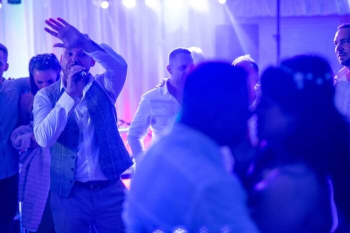 A táncparketten a násznép örömmel táncol. A háttérben a pár közelében, gyengén elmosódott fények között a vendégek éppen élvezik az este hangulatát. A fények és a színek segítenek megragadni a bulizós atmoszférát, miközben a fiatal házasok is együtt táncolnak, romantikus pillanatokat élve át.