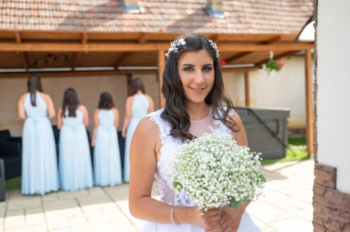 Egy menyasszony fehér ruhában, kezében apró fehér virágcsokorral mosolyog a kamera felé, miközben a koszorúslányok háttal állnak a háttérben, kék ruhában.