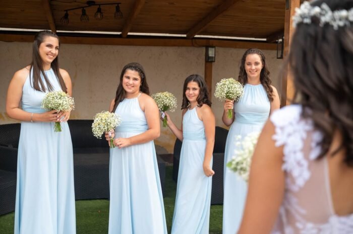 Koszorúslányok kék ruhában, kezükben fehér virágcsokorral mosolyognak, miközben a menyasszony alakja a háttérből látszik.