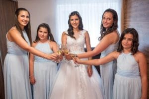 A menyasszony és koszorúslányai egy pohár pezsgővel koccintanak. A menyasszony fehér ruhát visel, míg a koszorúslányok világoskék ruhákban vannak. Az esemény egy világos szobában zajlik, természetes fényben.