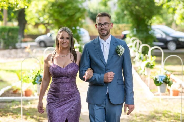 Az ifjú férj és édesanyja együtt sétálnak az esküvői helyszínre. Az ifjú férj kék öltönyt visel, virágcsokorral a zakóján, édesanyja pedig egy csillogó lila ruhában van. A háttérben az esküvői dekoráció látható, virágokkal díszítve az ösvényt.
