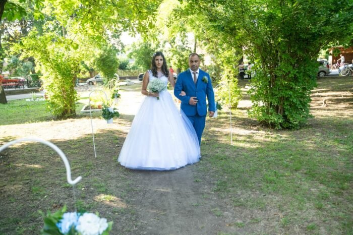 A menyasszony és édesapja a szabadtéri esküvői ceremóniára tartanak, miközben az esküvői szertartás helyszínéhez közelednek. A menyasszony csodálatos fehér ruhát visel, miközben virágcsokrot tart, édesapja pedig kék öltönyben kíséri őt. A háttérben az esküvői helyszín és a zöld növények díszítik a tájat.