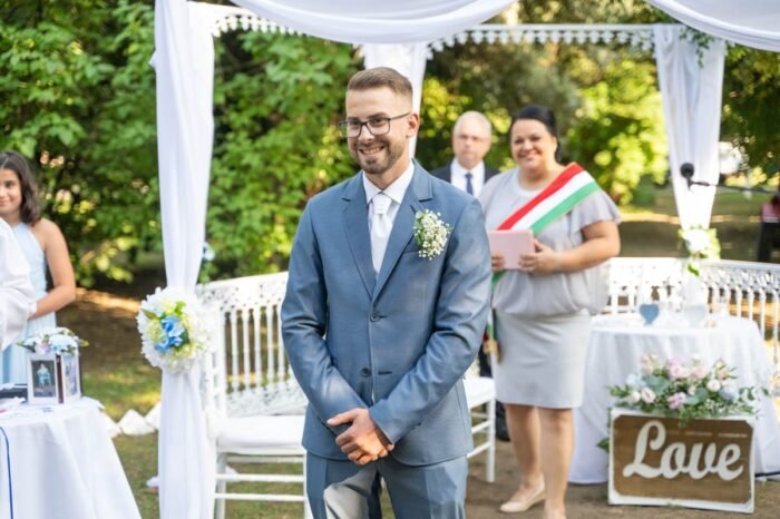 Az ifjú férj mosolygósan várakozik az esküvői szertartás alatt a szabadtéri helyszínen, miközben menyasszonya felé fordul. Az esküvői szertartást vezető személy éppen beszél, miközben az asztalon virágok és egy "Love" feliratú tábla is látható. A háttérben az esküvői vendégek és egy szép zöld környezet látszik.