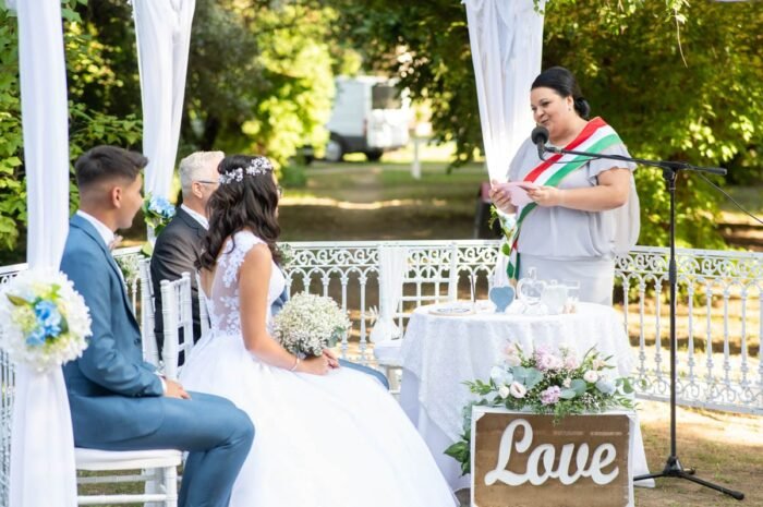 Az ifjú pár a szabadtéri esküvői szertartásán ül a házasságkötő asztal előtt, miközben az esküvői szertartást vezető személy beszél. A menyasszony kezében a virágcsokor, és ő figyelmesen hallgatja a beszédet. Az esküvői helyszínen egy szép "Love" feliratú tábla található, és a háttérben látszanak a násznép egyes tagjai is.