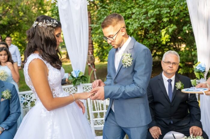 Az ifjú pár épp az esküvői szertartás közben cserél gyűrűt a szabadtéri oltár előtt. A menyasszony gyönyörű, csillogó esküvői ruhában áll, miközben vőlegénye elhelyezi az ékszert az ujján. A háttérben násznép figyeli őket, és a szertartást vezető hivatalos személy is jelen van. A képen a szeretet és az érzelem a házaspár arcán is tükröződik.