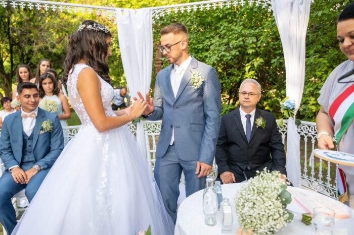 Az ifjú pár esküvői szertartás közben, az oltár előtt, ahol a menyasszony és a vőlegény épp a gyűrűket cserélik. A háttérben a násznép figyeli a szertartást, és az esküvőt vezető hivatalos személy is jelen van. A menyasszony gyönyörű esküvői ruhában, míg a vőlegény elegáns öltönyben áll, és az érzelmek mindkettőjük arcán tükröződnek.