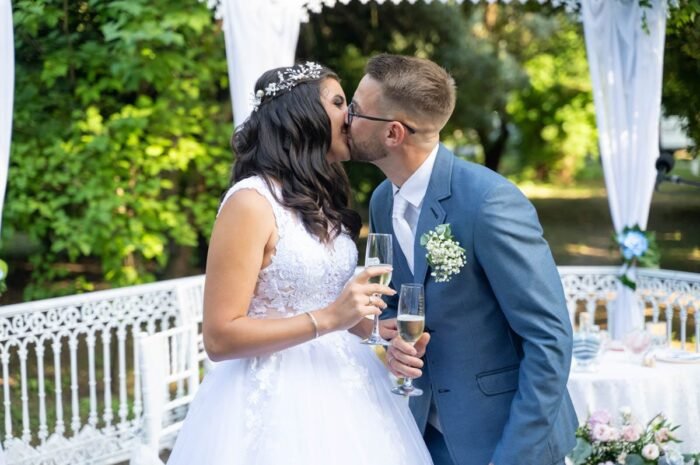 Az ifjú pár egymást csókolja meg esküvőjük napján, miközben pezsgőt tartanak a kezükben. A háttérben az esküvői díszítés, fehér textíliával és virágokkal, valamint a zöldellő fák adnak romantikus hangulatot a pillanathoz.