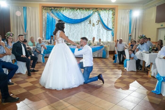 A vőlegény térdelve kezét nyújtja a menyasszonynak, miközben egy szép pillanatra figyelmezteti a násznépet. A háttérben az esküvői dekoráció és az ünneplő vendégek figyelnek, miközben a pár különleges pillanatot oszt meg.