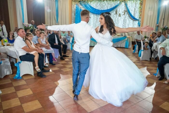 A menyasszony és a vőlegény lenyűgöző első táncukat járják, miközben a násznép figyelmesen nézi őket. A pár elegánsan pörög a táncparketten, miközben a menyasszony ruhája bájosan forog körülöttük. A háttérben a család és barátok élvezik az ünneplést.