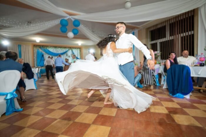 A menyasszony és a vőlegény vidáman táncolnak, miközben a menyegzői hangulat a háttérben folytatódik. A menyasszony tánc közben forgolódik, így a ruhája elegánsan lebeg körülötte. A násznép a háttérben élvezi az ünneplést, miközben a tánc örömteli pillanatait megörökítik.