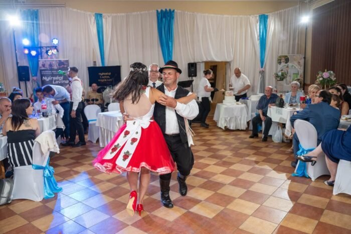 Egy menyasszony piros-fehér népi ruhában táncol egy férfival a menyegzőn, miközben a násznép az asztaloknál ül és figyeli őket. A háttérben a torta, a dekoráció és a fények láthatók. A tánc örömteli és hagyományos hangulatot áraszt, miközben a rendezvény folytatódik.