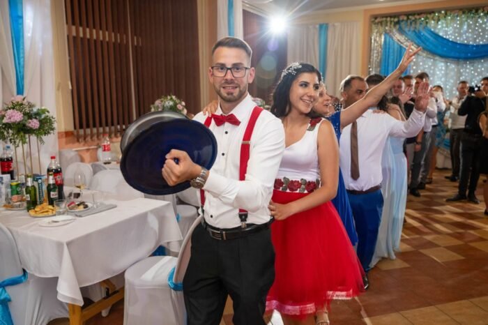 A menyasszony és vőlegény táncolnak, miközben a násznép ünnepel a háttérben. A menyasszony piros-fehér ruhában, a vőlegény piros masnival díszített fehér ingben és piros nadrágtartóban van. Az asztalon italok és harapnivalók láthatók, és a dekorációban kék és fehér színek dominálnak.