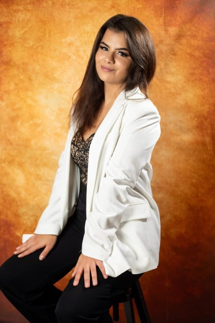 Fiatal nő portréja stúdiófotón, fehér zakóban és fekete csipkés felsőben. Háttérként meleg, aranyló tónusú színek szolgálnak, amelyek hangsúlyozzák a modell kifinomultságát és természetes szépségét.