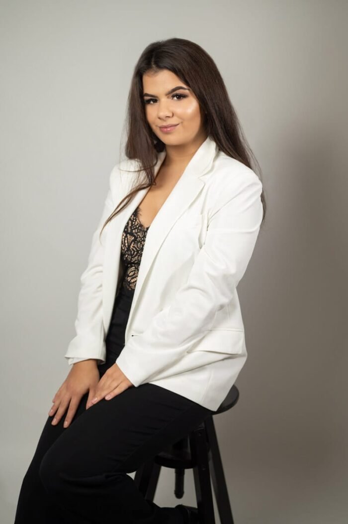 Portré fotózás Debrecenben. Fiatal nő elegáns stílusban, fehér zakóban és fekete csipkés felsőben, egy minimalista, világosszürke háttér előtt. Finom megjelenése és természetes mosolya magabiztosságot és stílust sugároz.