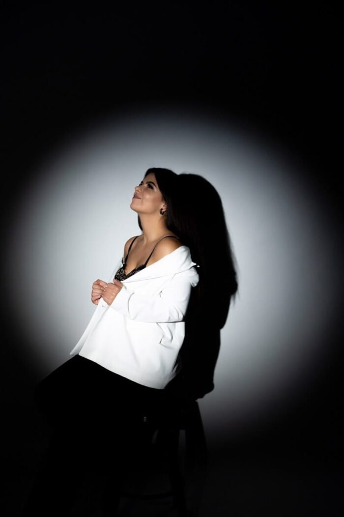 Fiatal nő, elegáns fehér zakóban és fekete csipkés felsőben, erőteljes fényfoltos megvilágításban. Az árnyékok játéka és a kontrasztos világítás lenyűgöző drámaiságot kölcsönöz a képnek, miközben a modell felfelé nézve inspirációt és reményt sugall.