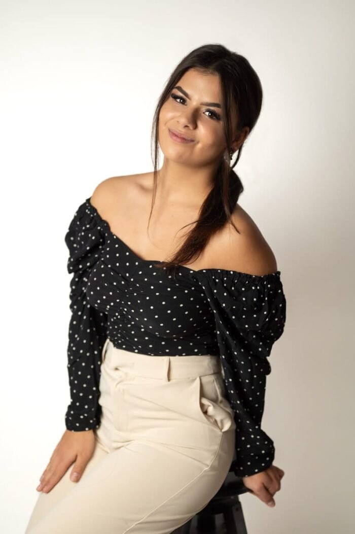 Fiatal nő világos háttér előtt, fekete-fehér pöttyös felsőben és bézs nadrágban ül. Mosolygós arckifejezése, oldalra húzott frizurája és egyszerű, letisztult stílusa friss és modern megjelenést sugall.