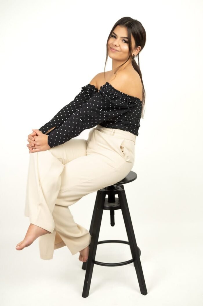 Fiatal nő fekete-fehér pöttyös felsőben és bézs nadrágban, mosolygósan ül egy magas széken. Letisztult, modern stílusa és természetes eleganciája Debrecen fotóstúdiójának minimalista hátterével párosul.