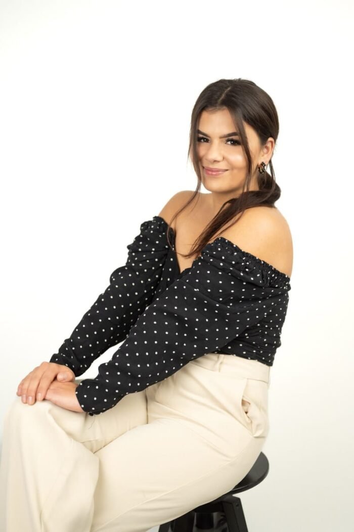 Fiatal nő fekete pöttyös, vállakat szabadon hagyó felsőben, bézs nadrágban mosolyog egy fotóstúdió minimalista fehér háttere előtt Debrecenben