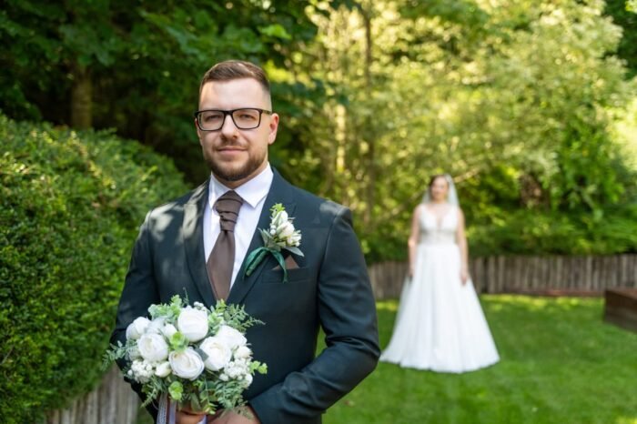 Vőlegény fekete öltönyben, fehér virágcsokrot tartva, előtérben áll, miközben a menyasszony fehér ruhában a háttérben mosolyog egy zöld kertben.