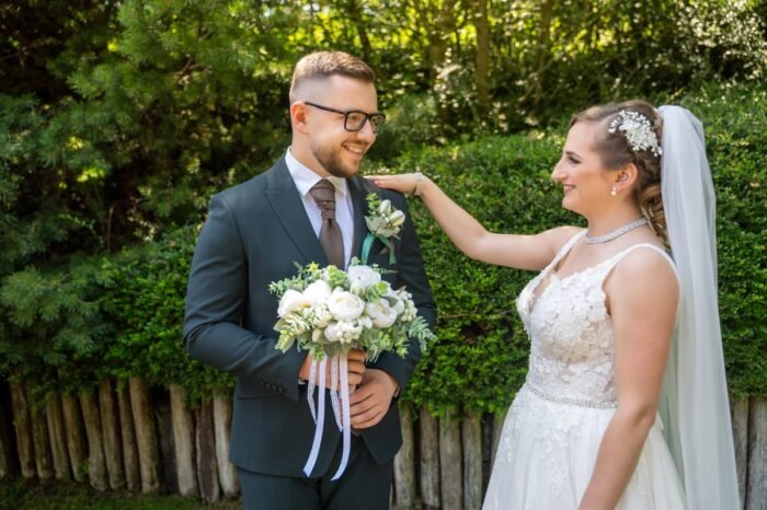 A vőlegény mosolyogva áll fekete öltönyben, virágcsokrot tartva, miközben a menyasszony fehér csipkeruhában gyengéden a vállára teszi a kezét egy zöld kertben.