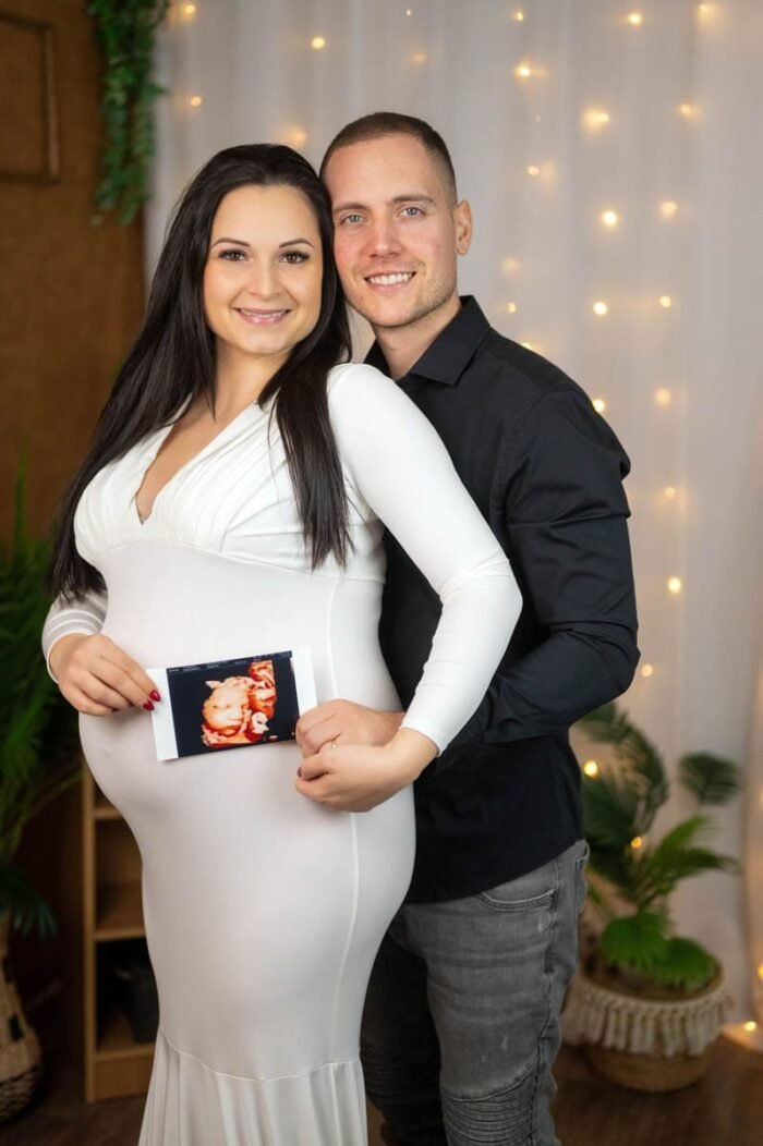 A pár boldogan áll egymás mellett, miközben a kismama a terhét tartja, és az ultrahangos képet a hasához helyezi. Az apa mögötte mosolyogva öleli át, mindketten örömmel mutatják a kisbabájuk képeit, miközben a háttérben fények és növények díszítik a hangulatos teret.