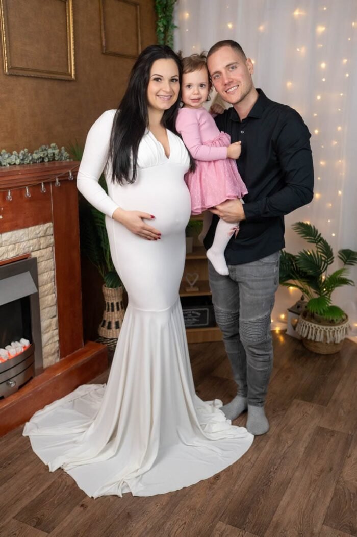 Családi kismama fotózás Debrecen. Egy boldog család: a kismama elegáns fehér ruhában, párja és kislányuk mosolyogva állnak egymás mellett egy ünnepi dekorációval díszített háttér előtt. A kislány rózsaszín ruhát visel, a meghitt hangulatot fényfüzérek és növények teszik teljessé.