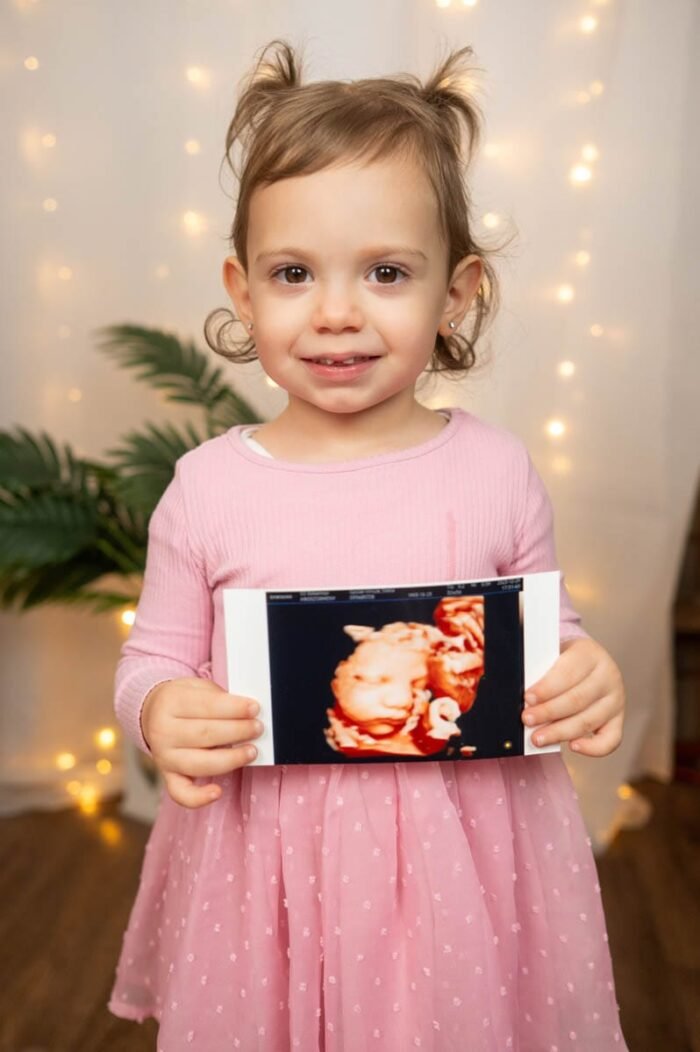 Gyermek fotózás Debreceni stúdióban. A kislány mosolyogva tartja testvére ultrahangos képét, miközben a háttérben meghitt fények világítanak. Az apró lány rózsaszín ruhában, kedvesen néz a kamerába, miközben az ultrahangkép egy kisbabát ábrázol, amit büszkén mutat a szüleinek.