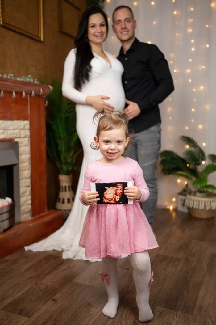 Családi kismama fotózás Debrecenben - Egy kislány rózsaszín ruhában boldogan tartja a testvére ultrahangképét, miközben a háttérben a szülei szeretetteljes mosollyal állnak. Az anyuka fehér ruhában kiemeli pocakját, míg az apuka gyengéden mellette áll. A meghitt pillanatot hangulatos fények és természetes dekorációk teszik még különlegesebbé.