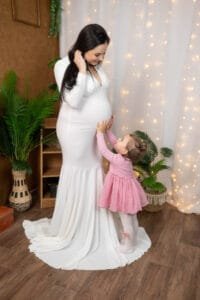 Egy kismama hófehér ruhában szeretettel mosolyog a kislányára, aki gyengéden megérinti az anyukája pocakját. A háttérben meleg fények és növények díszítik a meghitt jelenetet, a kislány rózsaszín ruhájában boldogan néz anyukájára, tele szeretettel és kíváncsisággal.