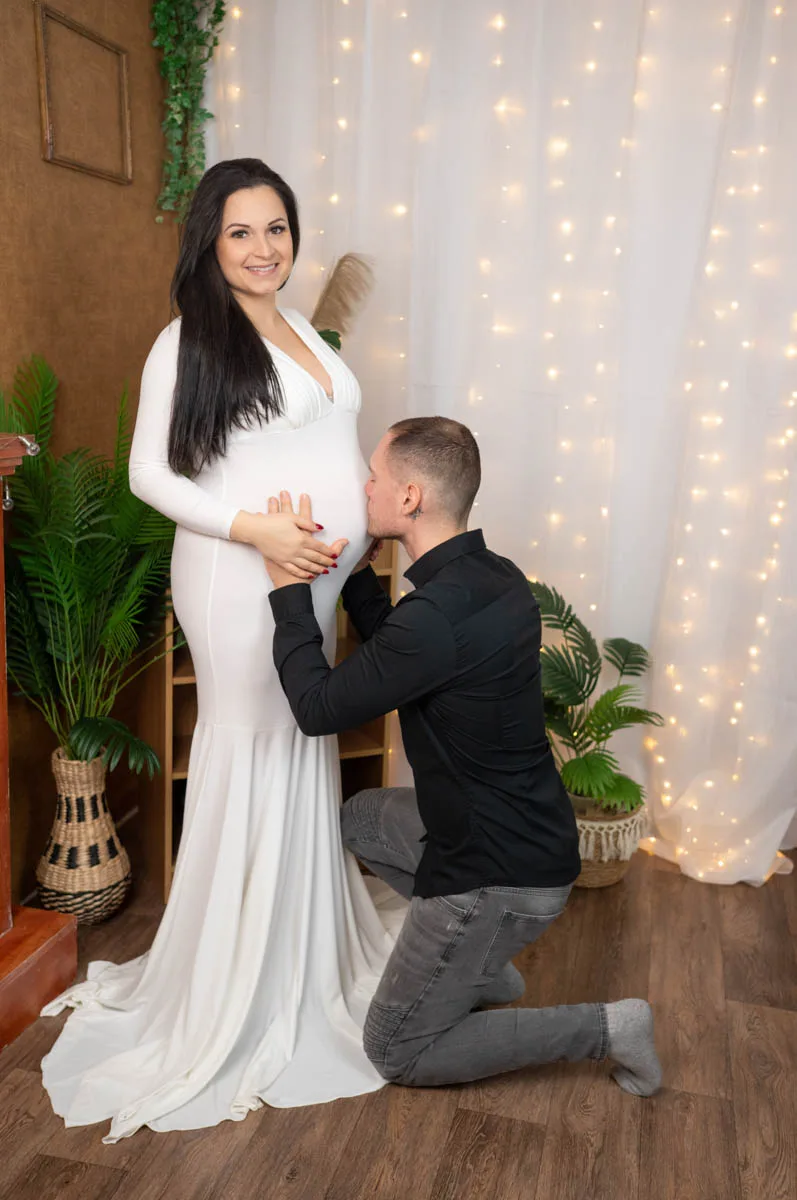 Kismama fotózás Debrecenben- Egy mosolygó kismama hosszú fehér ruhában, aki boldogan áll a férje előtt, miközben ő szeretetteljesen megpuszilja a pocakját.