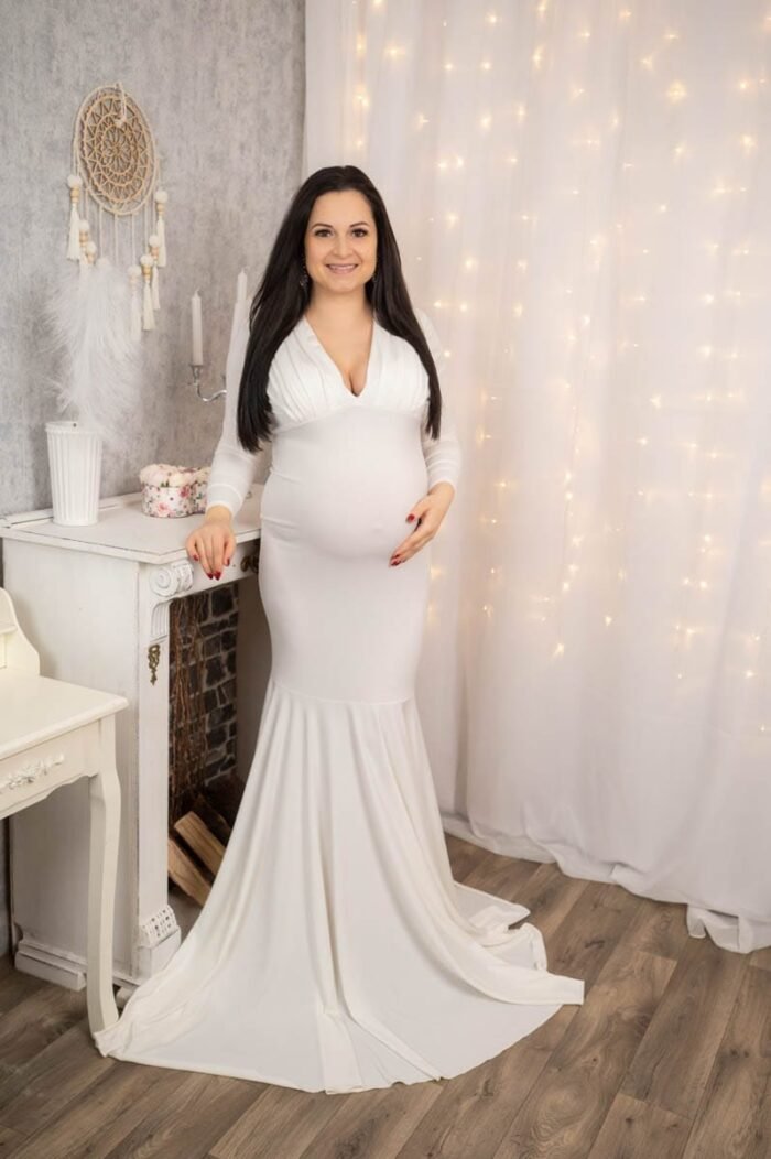 Egy elegáns kismama hosszú, fehér ruhában, vintage stílusú bútorokkal és fényfüzérrel díszített meghitt háttér előtt. A kép harmonikus és ünnepi hangulatot áraszt, kiemelve az anyaság szépségét és örömét.