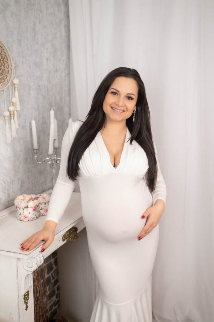 Egy mosolygó kismama hosszú, fehér ruhában, vintage stílusú, virágokkal és gyertyatartókkal díszített háttér előtt. Az egyszerű, letisztult kompozíció kiemeli a kismama természetes szépségét és az anyaság meghitt pillanatát.