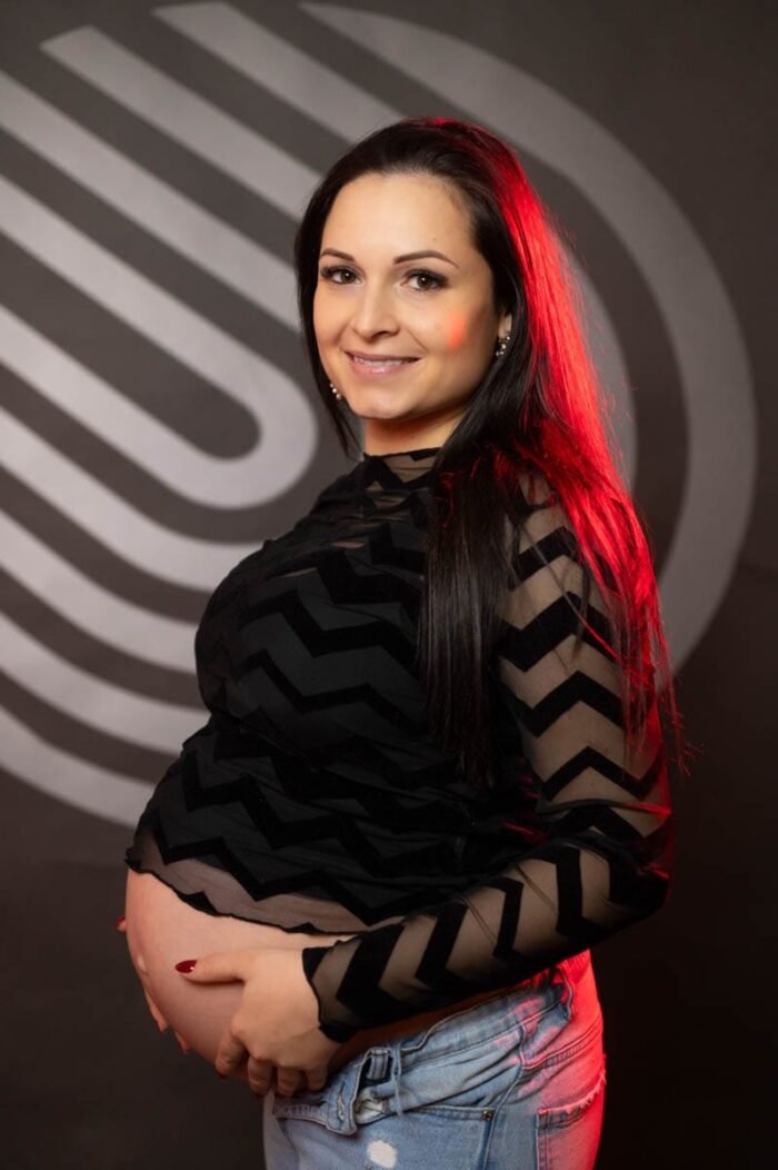 Egy mosolygós kismama modern stílusú háttér előtt. A fekete mintás felső és a kék farmer kiemelik a divatos megjelenését, miközben gyengéden tartja pocakját. A háttér geometrikus mintái és a vörös fényhatás különleges hangulatot kölcsönöznek a képnek.