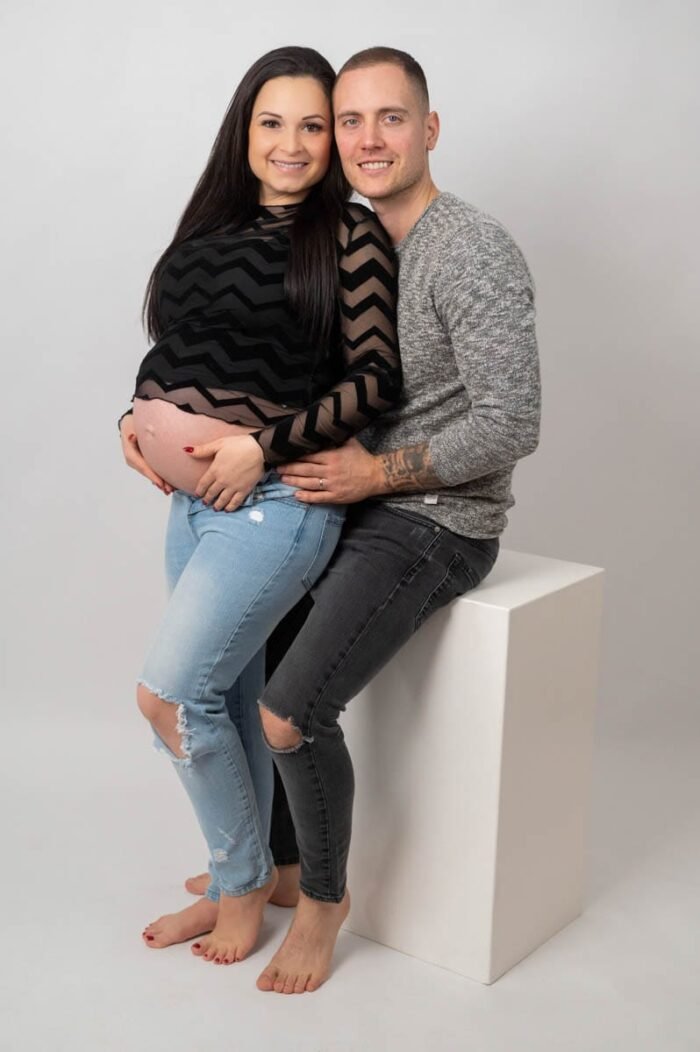 Egy mosolygós kismama és párja ülnek egy fehér kockánál kismama fotózáson. Az anya fekete mintás felsőt és világoskék farmert visel, míg párja szürke pulóverben öleli őt. Az anya pocakját mindketten gyengéden tartják, a szeretet és az összetartozás meghitt pillanatait közvetítve.