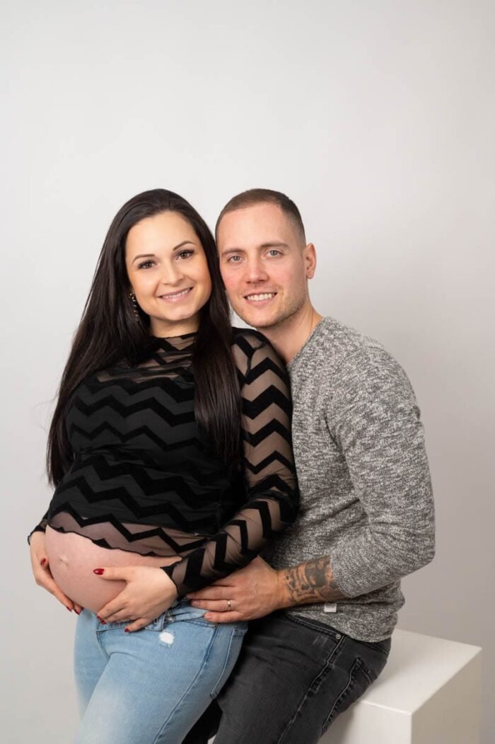 Egy boldog pár kismama fotózáson. Az anya fekete mintás felsőt és farmert visel, kezét a pocakján tartja, míg párja szürke pulóverben szeretetteljesen öleli őt. A háttér letisztult, a fókusz a pár örömén és a születendő gyermekük várakozásán van.