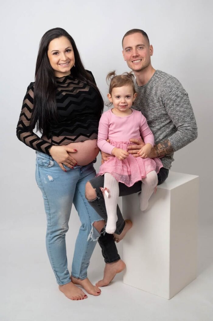 Egy boldog családot ábrázoló kép, ahol a kismama fekete mintás felsőben és farmerben áll, mellette párja ül a kislányukkal, aki rózsaszín ruhában mosolyog. A családi meghittség és az új élet érkezésének öröme sugárzik a képről.