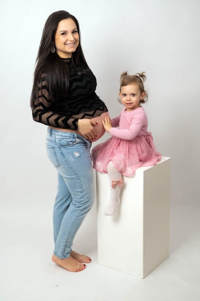 Egy kismama fekete mintás felsőben és farmerben áll, miközben kislánya rózsaszín ruhában ül egy fehér oszlopon, és kezét szeretetteljesen a kismama pocakjára helyezi. A világos háttér egyszerűsége kiemeli a családi kötődést és az érzelmi pillanatot.