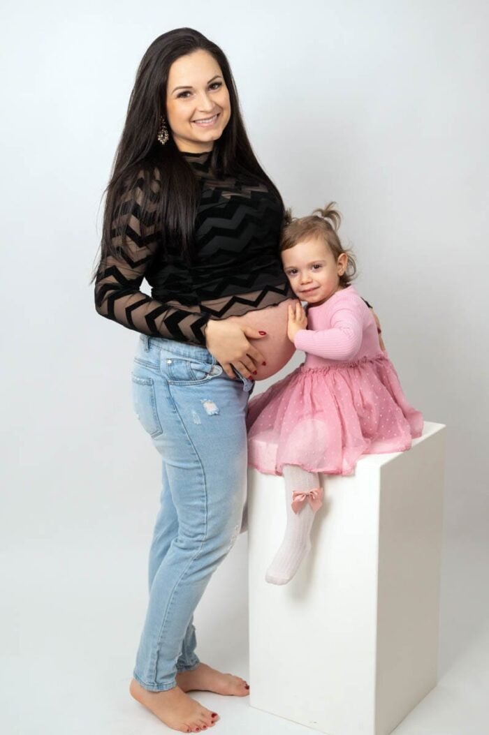 Egy kismama fekete mintás felsőben és farmerben áll, miközben kisgyermeke rózsaszín ruhában ül mellette egy fehér oszlopon. A kislány szeretetteljesen a kismama pocakjára helyezi a kezét, mosollyal az arcán, világos háttér előtt.