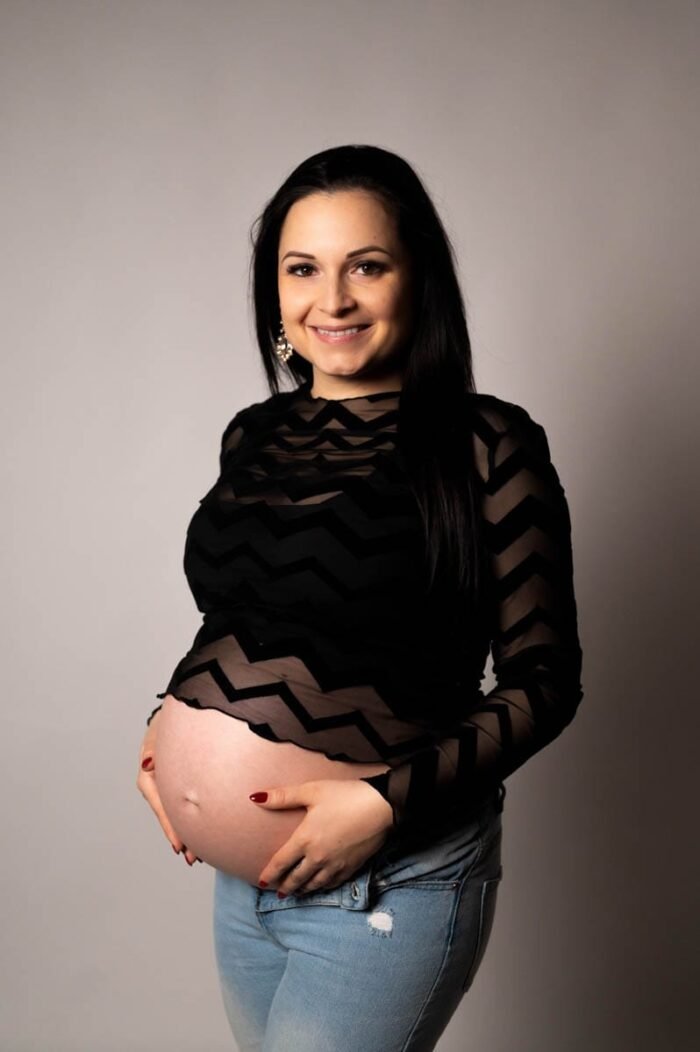 Kismama fotózás Hajdúböszörményben - Kismama fekete, áttetsző mintás felsőben és farmernadrágban, pocakját tartva, mosolyogva egy semleges szürke háttér előtt.