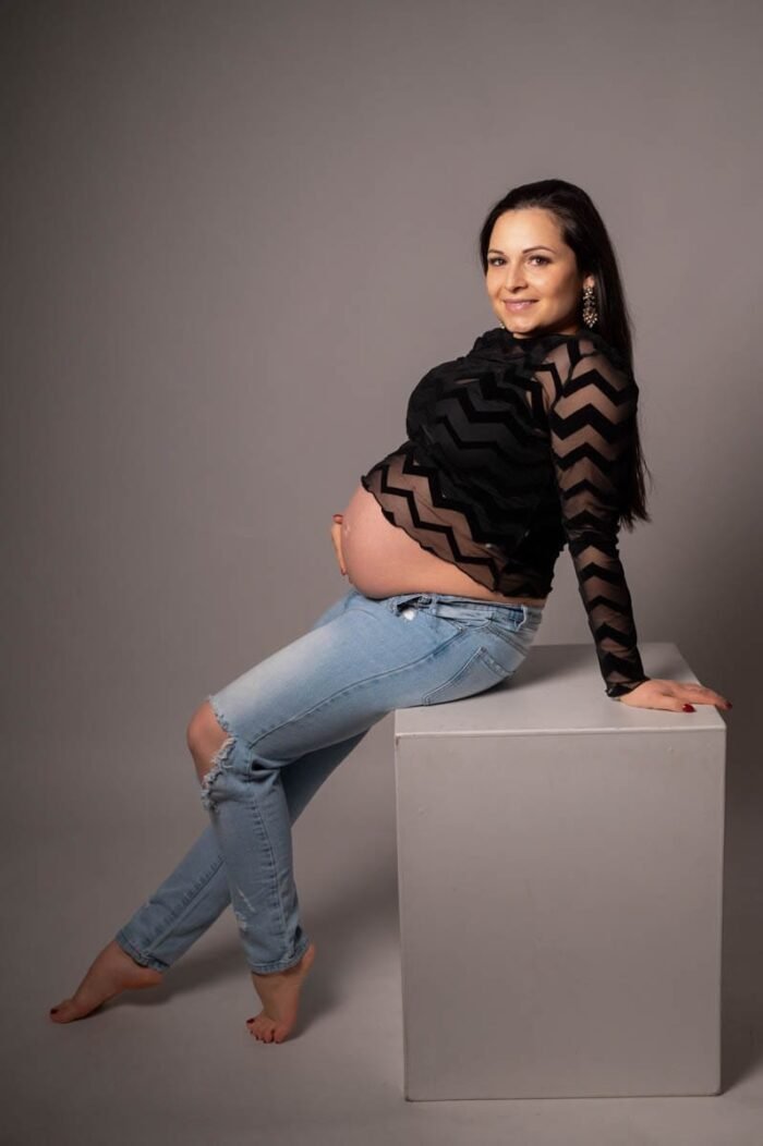 Várandós fotózás Debrecenben. Várandós nő farmerben és áttetsző fekete felsőben, mosolyogva ül egy fehér kockaformán, szürke stúdióháttér előtt.
