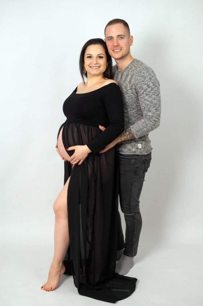 Egy mosolygó várandós nő fekete ruhában, mellette párja áll, szeretetteljesen átölelve őt. Fehér háttér előtt készült stúdiófotó.