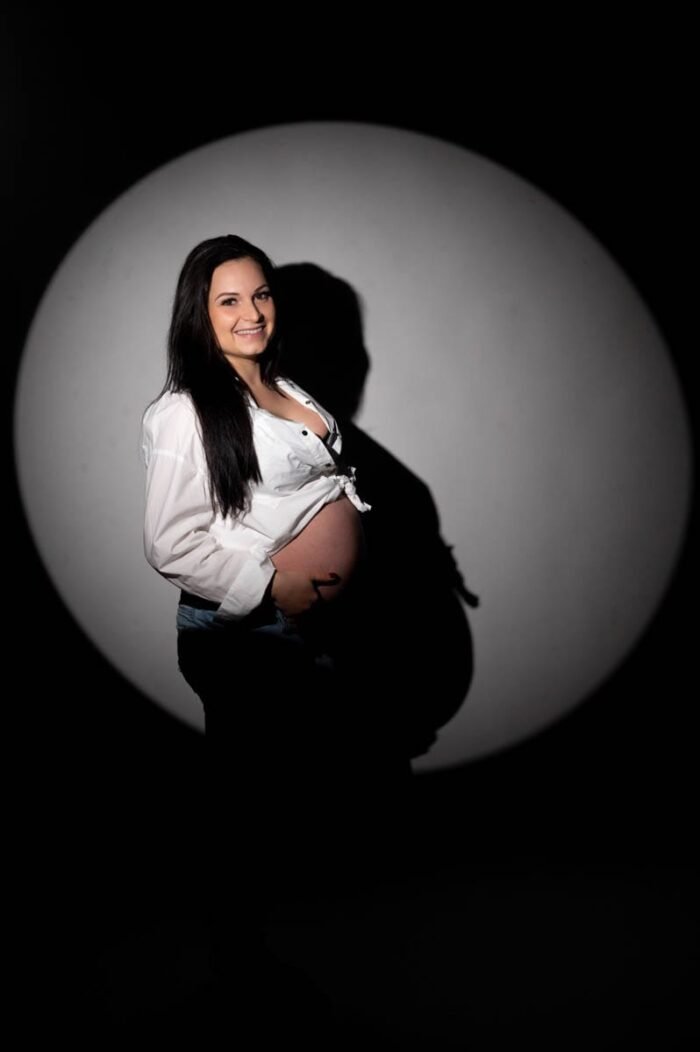 Egy várandós nő fehér ingben és farmerben áll, mosolyogva néz a kamerába. Sötét háttér előtt egy kör alakú fénykör világítja meg őt, árnyékát a falra vetítve.