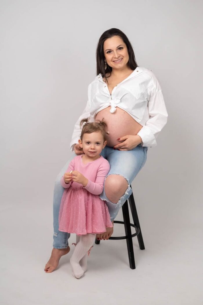 Egy várandós anya farmernadrágban és fehér ingben ül egy széken, mosolyogva pózol. Előtte kislánya rózsaszín ruhában áll, vidám arckifejezéssel, fehér háttér előtt.