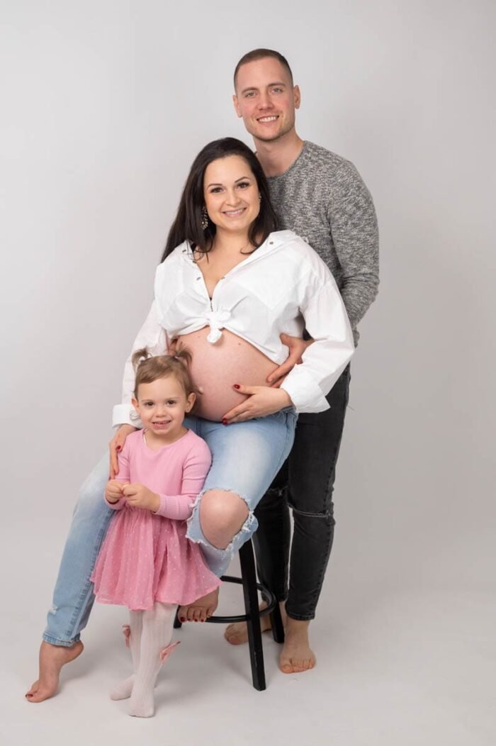 Egy boldog család pózol fehér háttér előtt. A várandós anya farmerben és fehér ingben ül egy széken, miközben férje átöleli és a pocakjára helyezi kezét. Előttük kislányuk áll rózsaszín ruhában, mosolyogva.