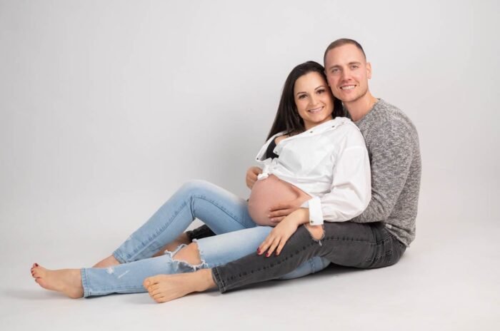 Pocakos fotózás Debrecen. Egy boldog pár ül fehér háttér előtt. A várandós nő farmernadrágban és fehér ingben mosolyog, miközben párja átöleli és a pocakjára helyezi kezét.
