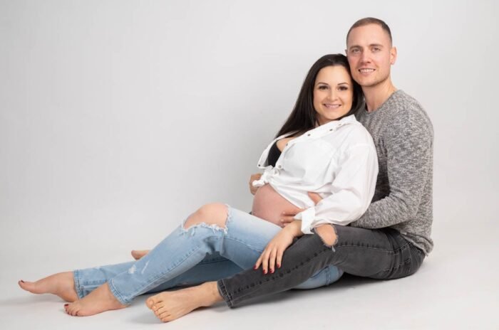 Egy mosolygó kismama és párja ülnek a fehér háttér előtt. A nő lezser fehér inget és farmernadrágot visel, kiemelve várandós hasát, miközben párja szeretettel átöleli.