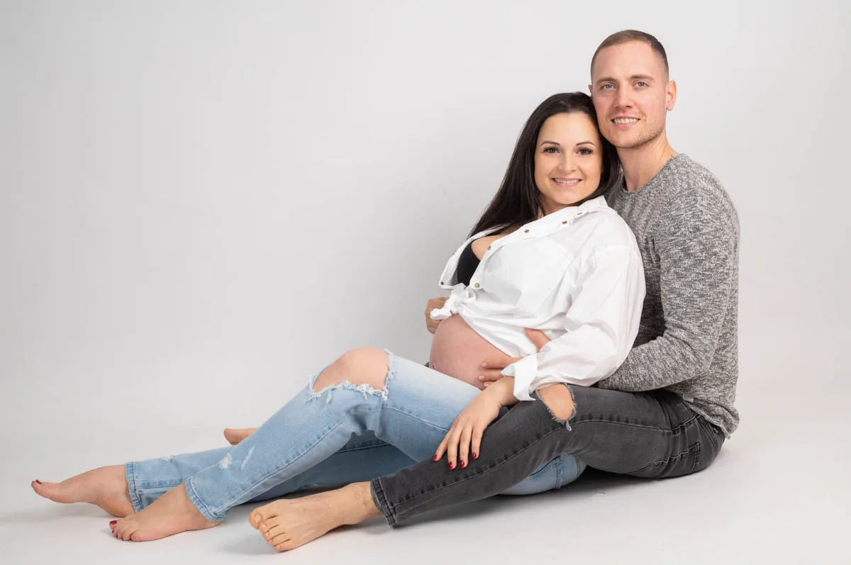 Egy mosolygó kismama és párja ülnek a fehér háttér előtt. A nő lezser fehér inget és farmernadrágot visel, kiemelve várandós hasát, miközben párja szeretettel átöleli.
